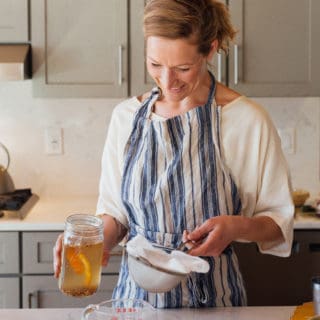A woman with blonde hair in a kitchen, wearing a white shirt and blue and white striped apron. She's holding a mason jar with liquid in one hand and a strainer with cheesecloth in the other.