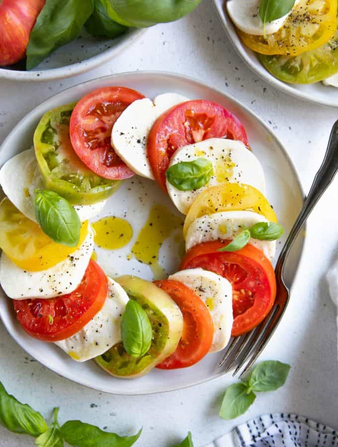 Slices of different colored tomatoes, mozzarella cheese and basil on a white plate with salt, pepper, and oiled drizzled on it.