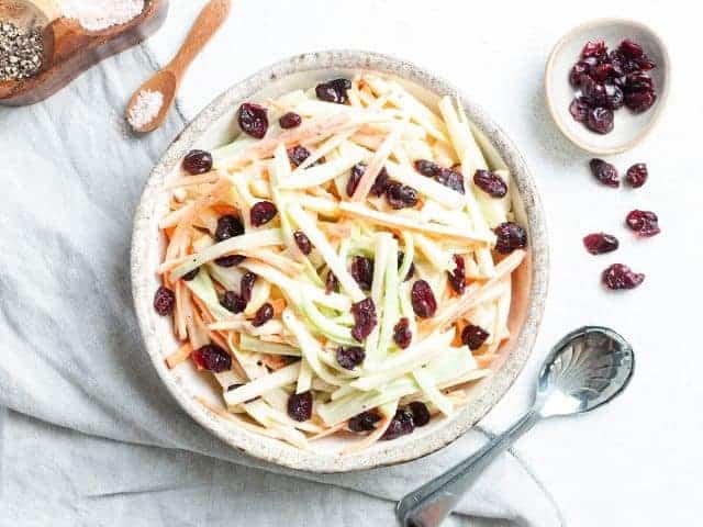 Shredded cabbage and carrots with raisins and a creamy dressing in a gray bowl and a metal spoon and other raisins next to it.