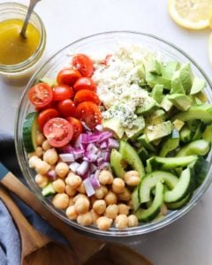 Top down shot of a large clear bowl with sliced cherry tomatoes, cucumber, chickpeas, chopped red onion, avocado, and feta cheese.