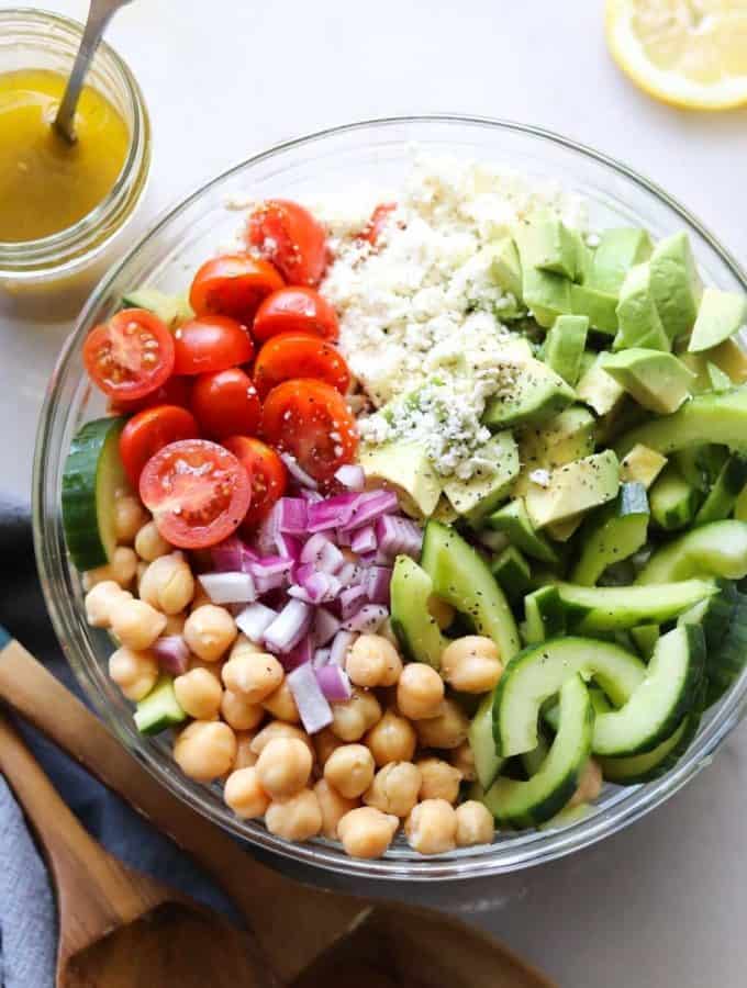 Top down shot of a large clear bowl with sliced cherry tomatoes, cucumber, chickpeas, chopped red onion, avocado, and feta cheese.