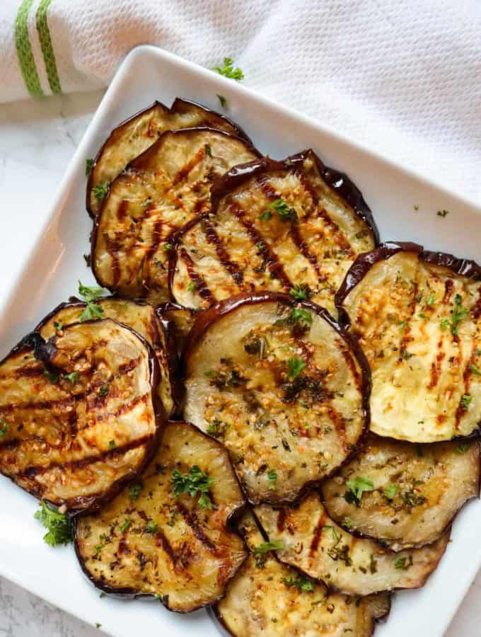 Slices of grilled eggplant on a square white platter next to a white and green towel.