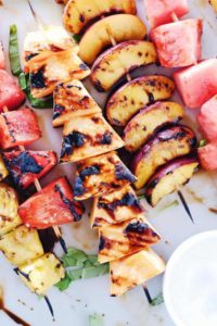 Pieces of grilled watermelon, peaches, and cantaloupe on wooden skewers lying on a white platter.