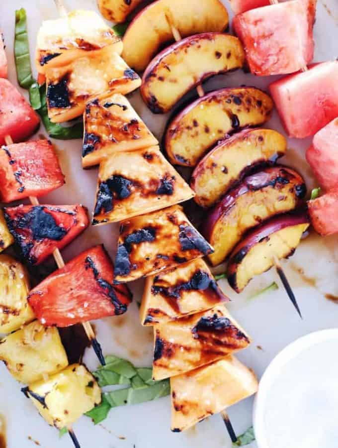 Pieces of grilled watermelon, peaches, and cantaloupe on wooden skewers lying on a white platter.