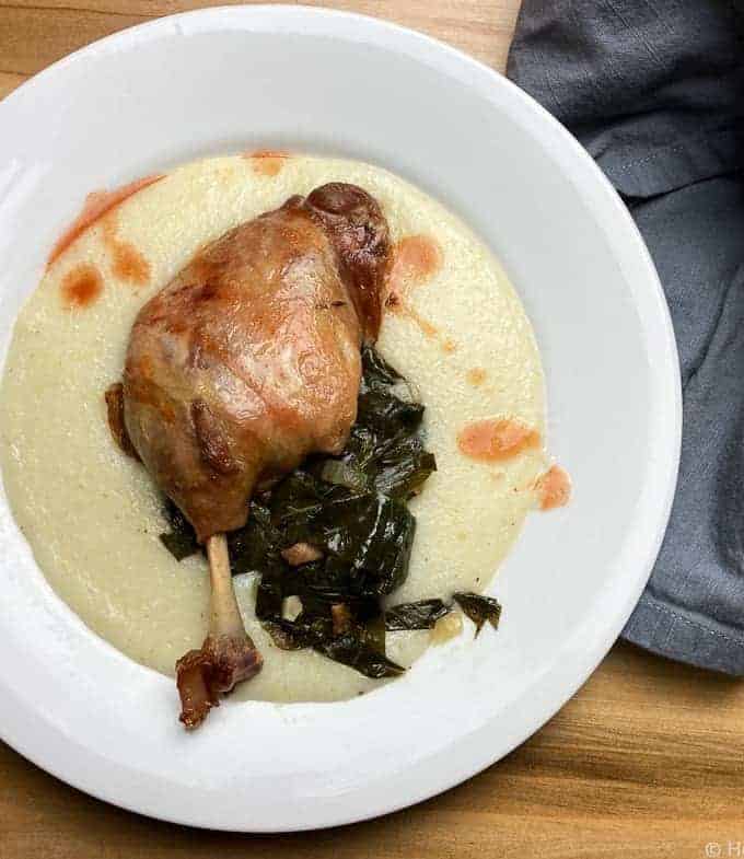 Top down shot of a white plate with grits, duck confit, and a cooked green leafy vegetable in it.