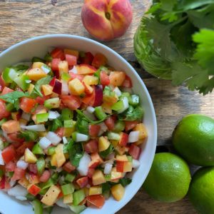 Top down shot of a peach salsa in a white bowl with limes, a peach, and fresh herbs to the side.