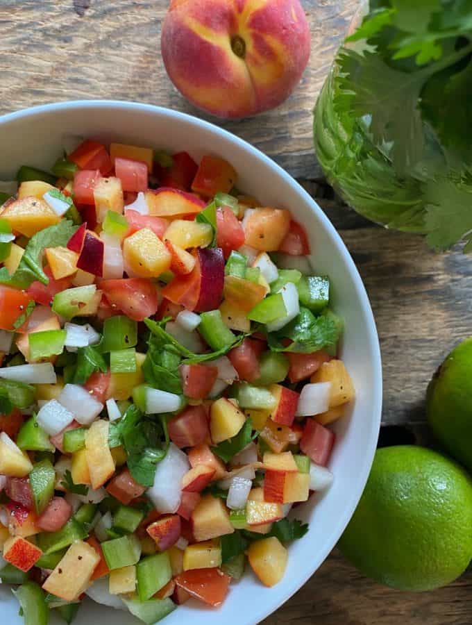 Top down shot of a peach salsa in a white bowl with limes, a peach, and fresh herbs to the side.