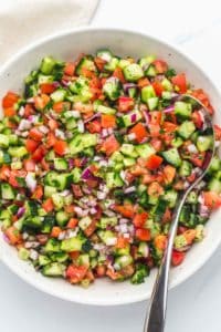 Diced cucumber, tomato, and purple onion with herbs and oil in a large white bowl with a metal serving spoon.