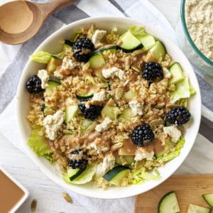 A salad of blackberries, zucchini, feta, quinoa, and lettuce in a white bowl with a brown dressing drizzled on top.