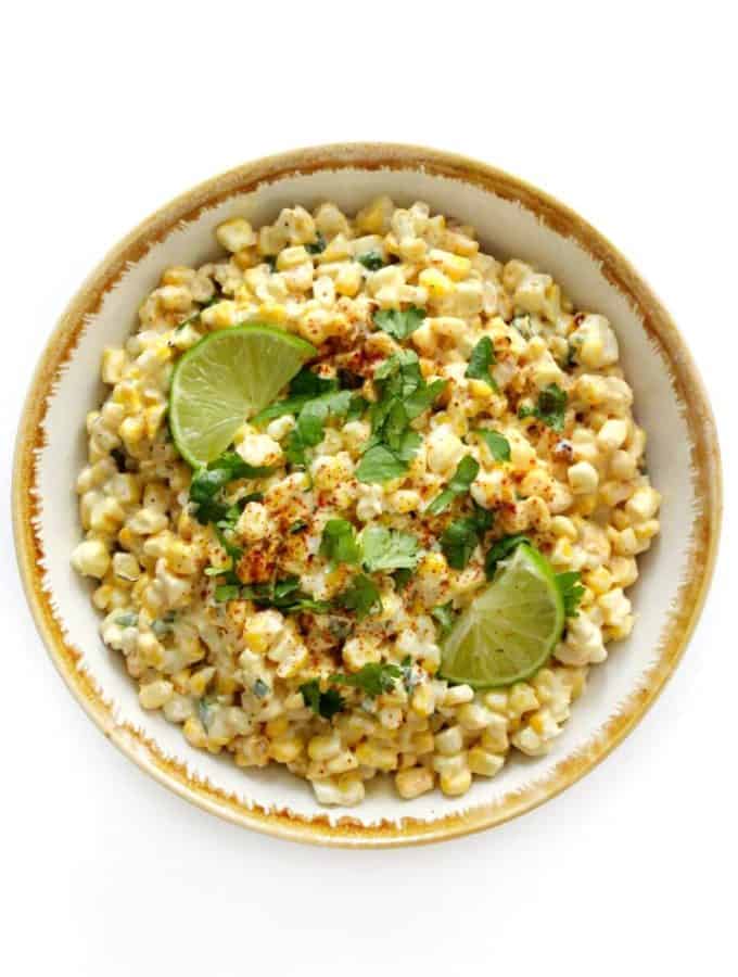 Top down shot of a corn salad in a white bowl topped with fresh herbs and a dash of a red spice.
