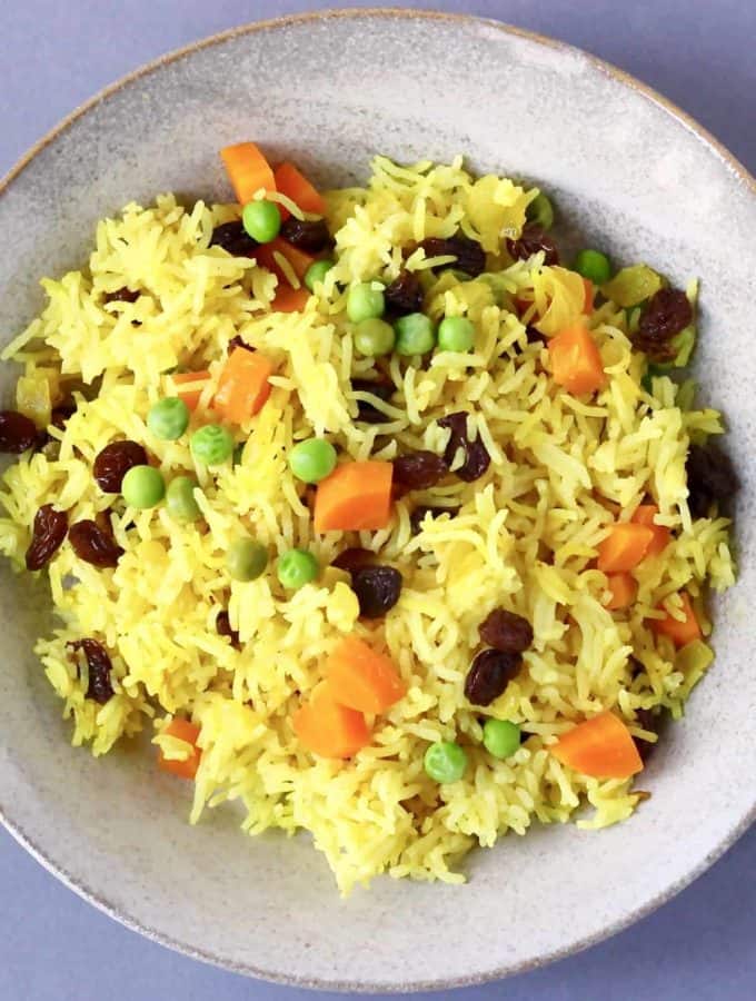 Top down shot of yellow rice with raisin, peas, and carrots in it on a gray plate.