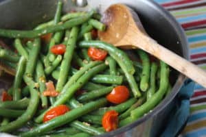 Close up of green beans and cherry tomatoes in a metal dish with a wooden spoon.