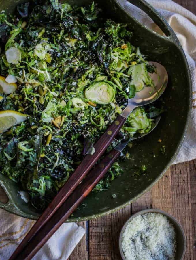 Shredded kale and Brussels sprouts with lemon slices in a large dark green bowl with serving utensils.