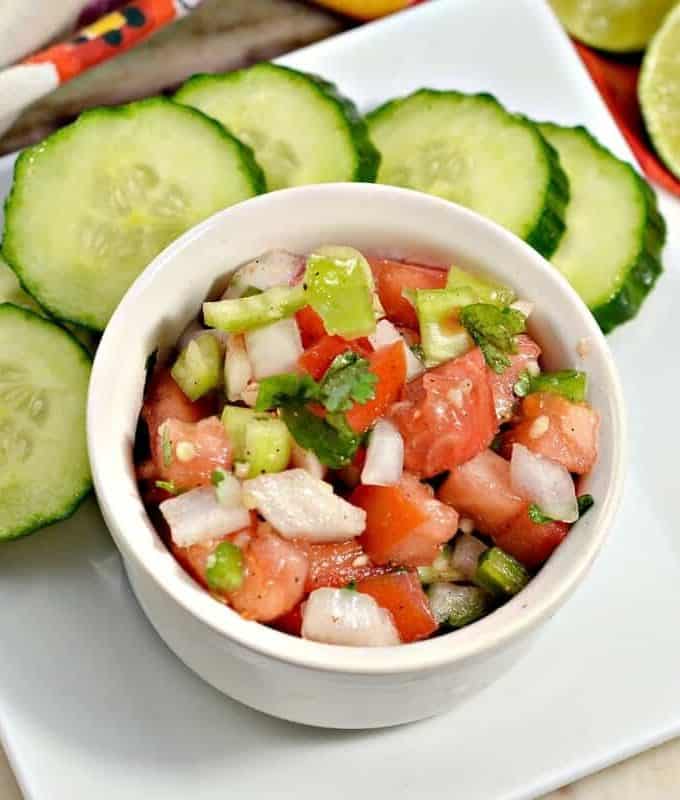 Close up of a pico de gallo in a small white round container with cucumber slices and limes next to it.