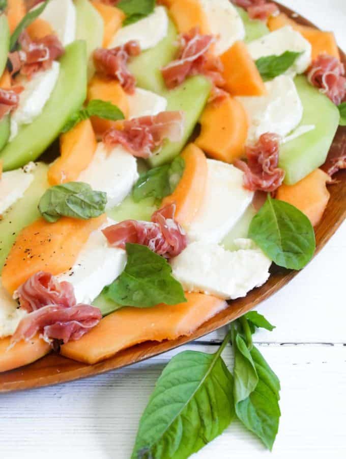 Slices of green and orange melon with mozzarella cheese, prosciutto, and basil on a wood platter.
