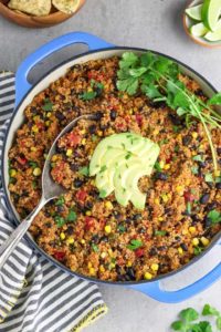 Top down shot of a red quinoa dish with corn, black beans, and tomatoes in it. Fresh herbs and sliced avocado are on top and it's in a blue serving pan.