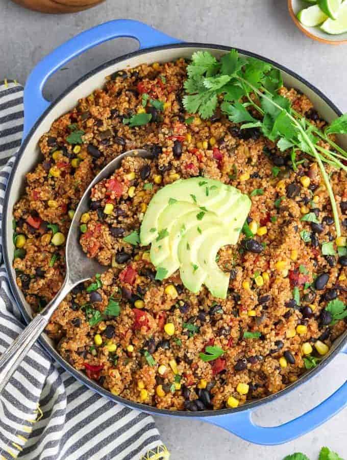 Top down shot of a red quinoa dish with corn, black beans, and tomatoes in it. Fresh herbs and sliced avocado are on top and it's in a blue serving pan.
