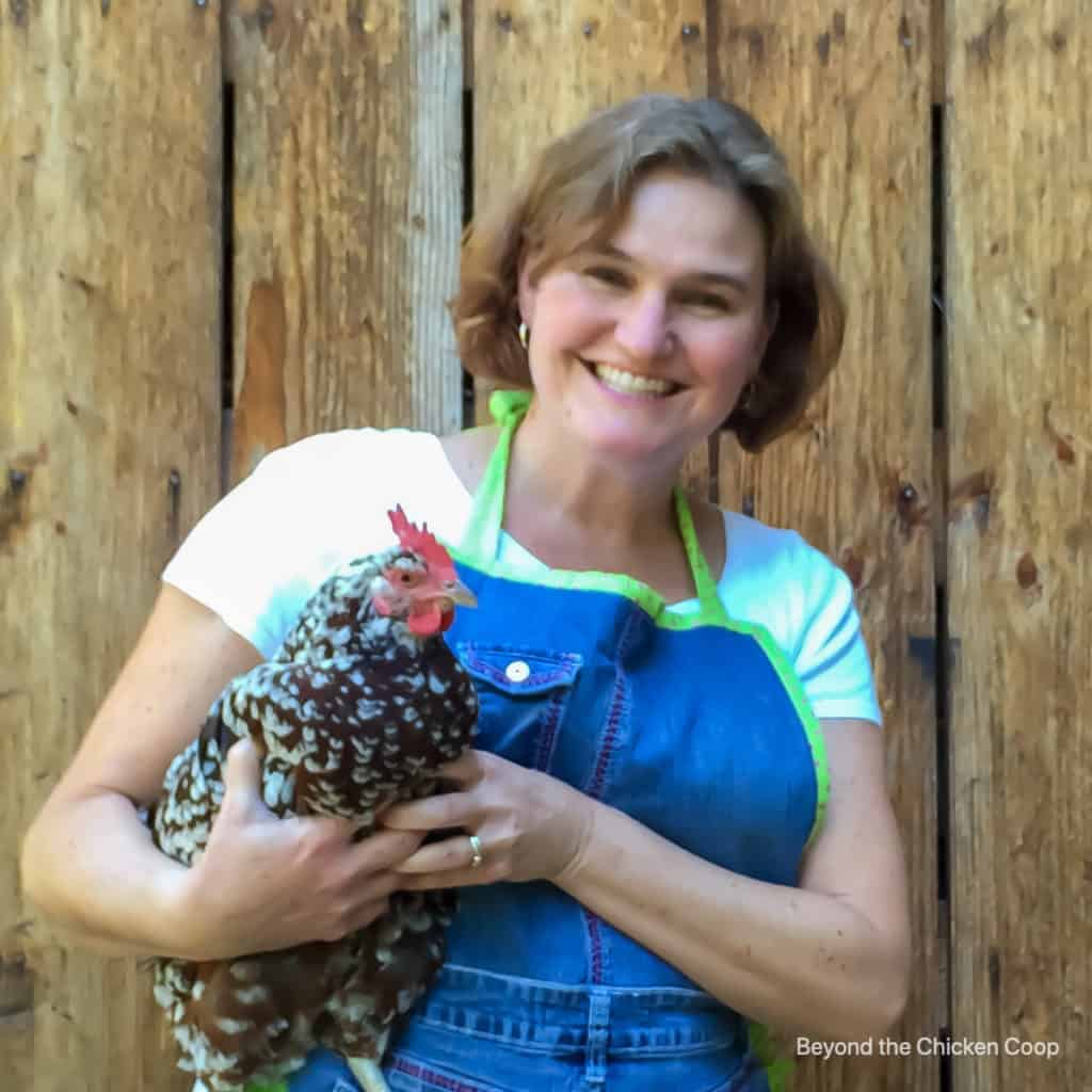 a woman with short brownish hard in a white t-shirt and blue apron holding a chicken in front of a wooden fence.