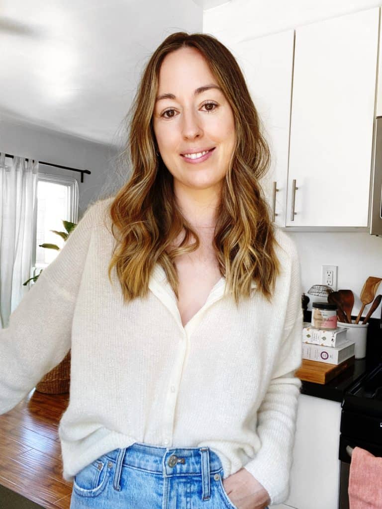 A tall thin woman with long brown hair in a off white button shirt and light blue jeans standing in a white kitchen.
