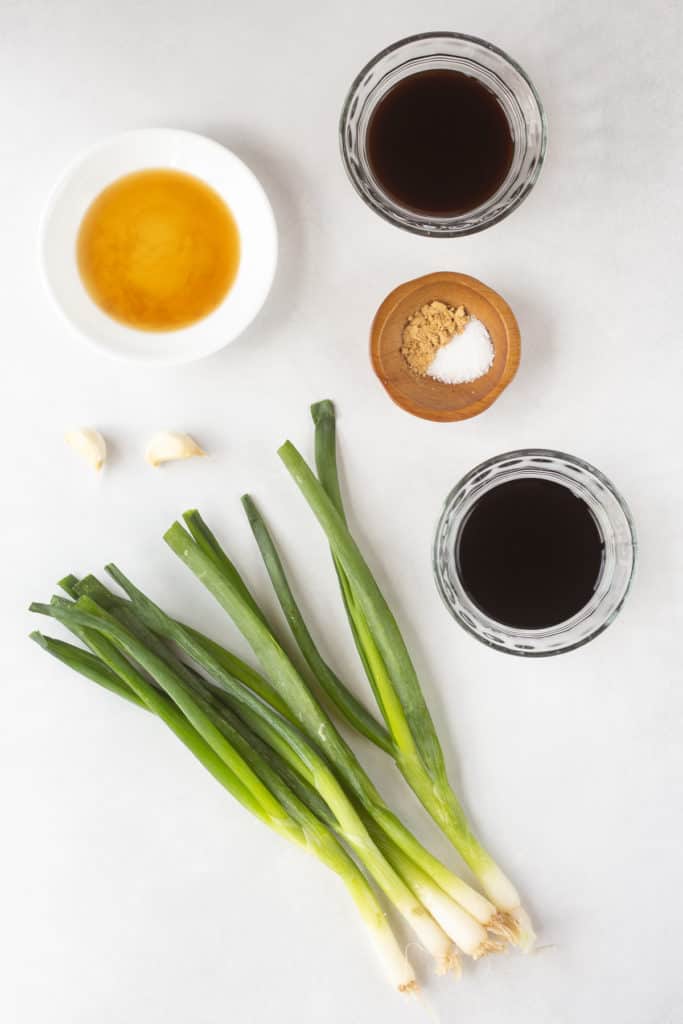 Top down shot of ingredients for a teriyaki marinade in clear and white bowls, as well as two cloves of garlic and five fresh green onions.