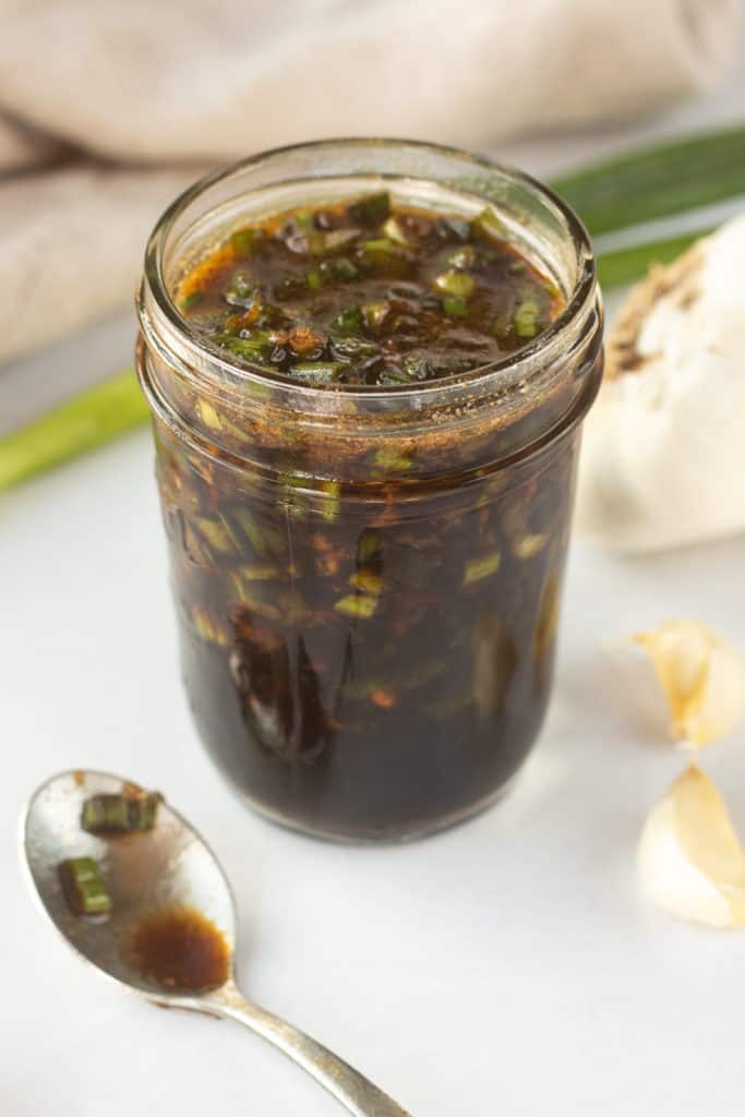 An angled shot of a mason jar with a brown marinade with chopped green onions in it with a small dirty spoon to the left and fresh green onions and cloves of garlic surrounding it.
