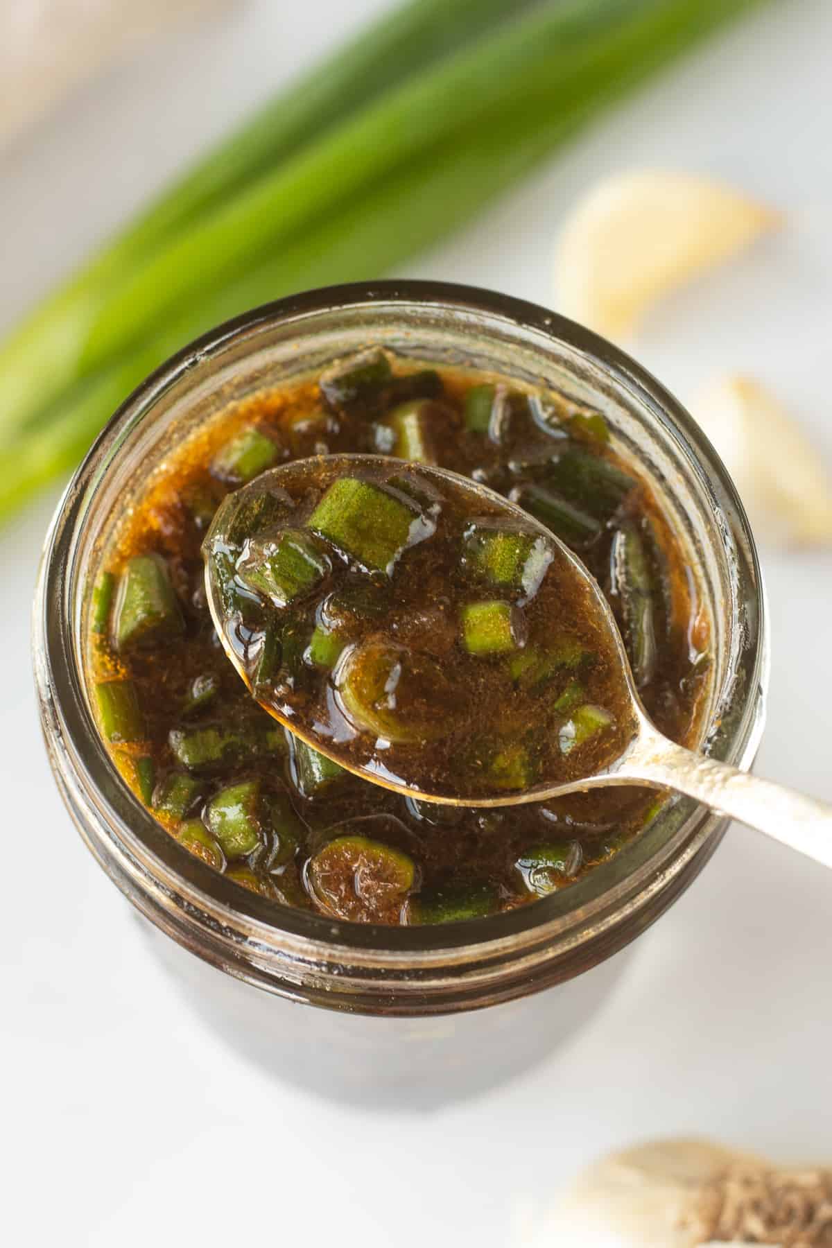 A small spoon dipping into a small mason jar with a brown marinade with chopped green onions in it. Fresh green onions and cloves of garlic are around the jar.
