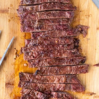 Top down shot of sliced skirt steak on a wood cutting board with a large knife and meat fork to the sides.