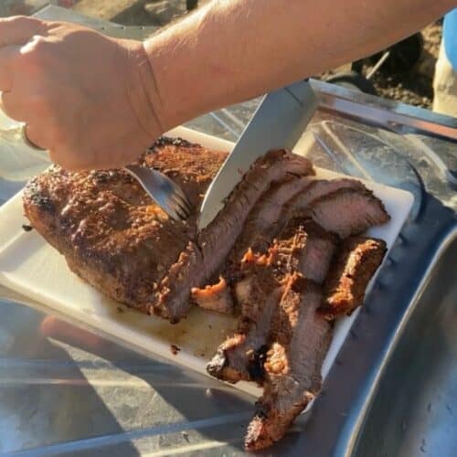 Two hands slicing grilled flank steak on a white cutting board in the setting sun.