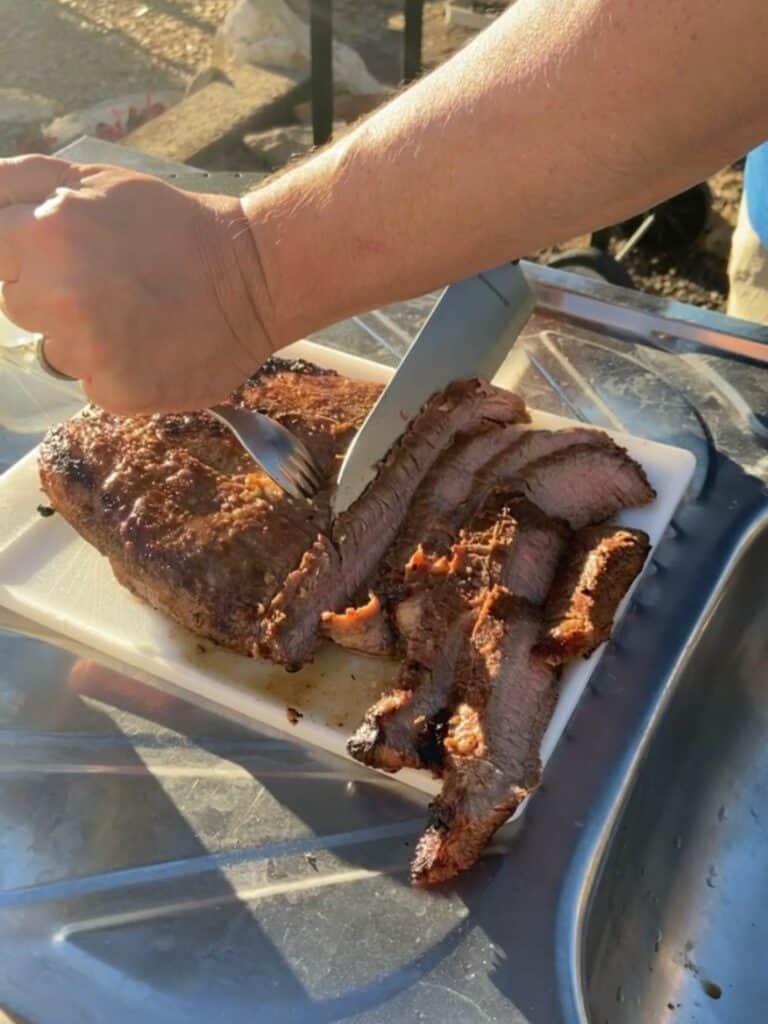 Two hands slicing grilled flank steak on a white cutting board in the setting sun.
