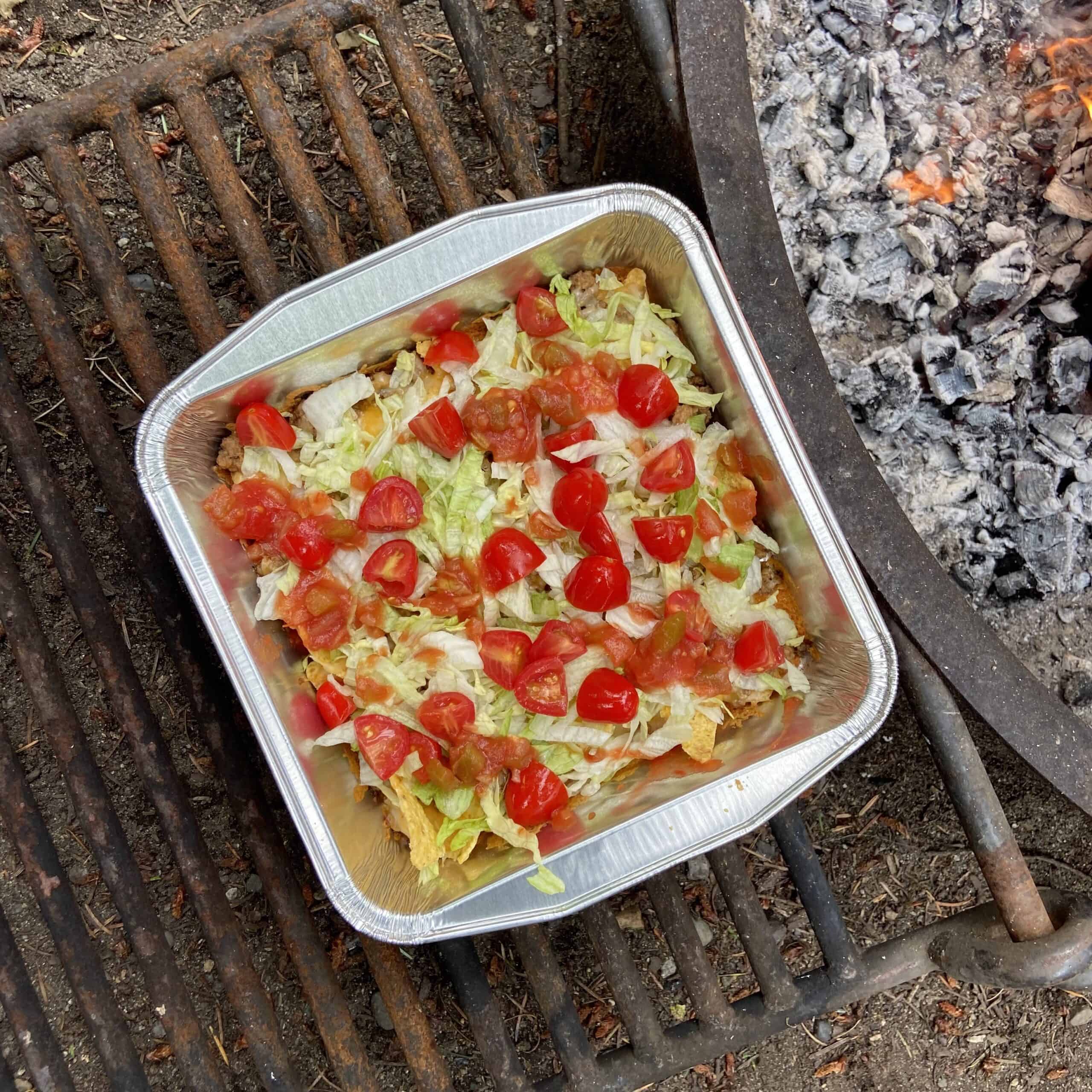 Top down shot of an aluminum 8x8 dish with nachos covered in lettuce and tomatoes in it. It's resting on a fire pit grate that's flipped over next to a fire pit.