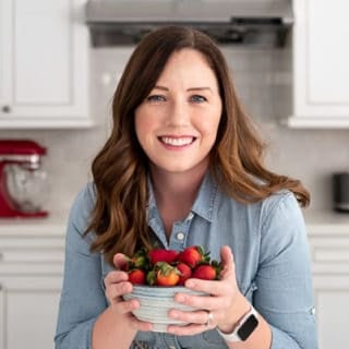 A middle-aged woman with long brown hair in a blue button up shirt holding a bowl of strawberries in a white kitchen.