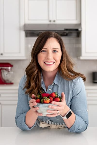 A middle-aged woman with long brown hair in a blue button up shirt holding a bowl of strawberries in a white kitchen.