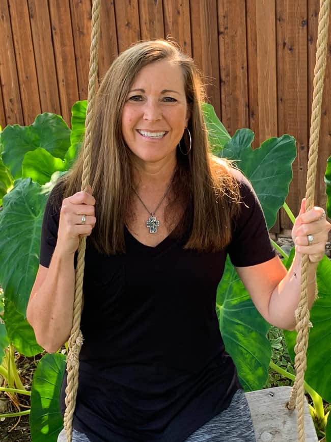 A middle aged woman with long brown hair in a black v-neck t-shirt sitting on a swing in front of a wooden fence.
