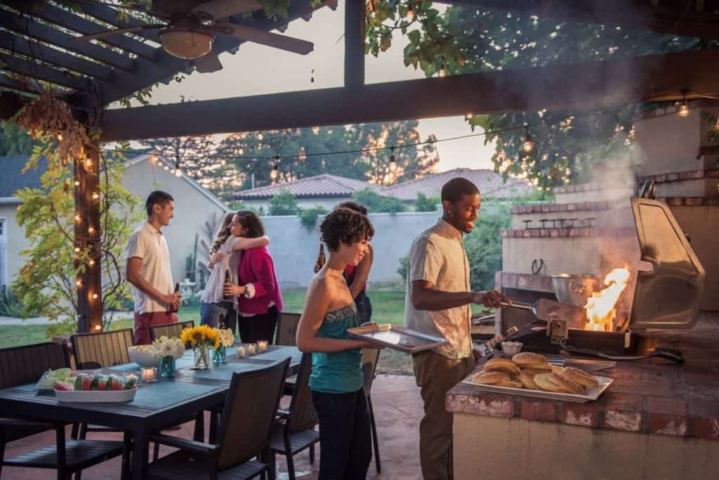 landscape shot of a crowd of younger adults outdoors around a table and grill socializing.