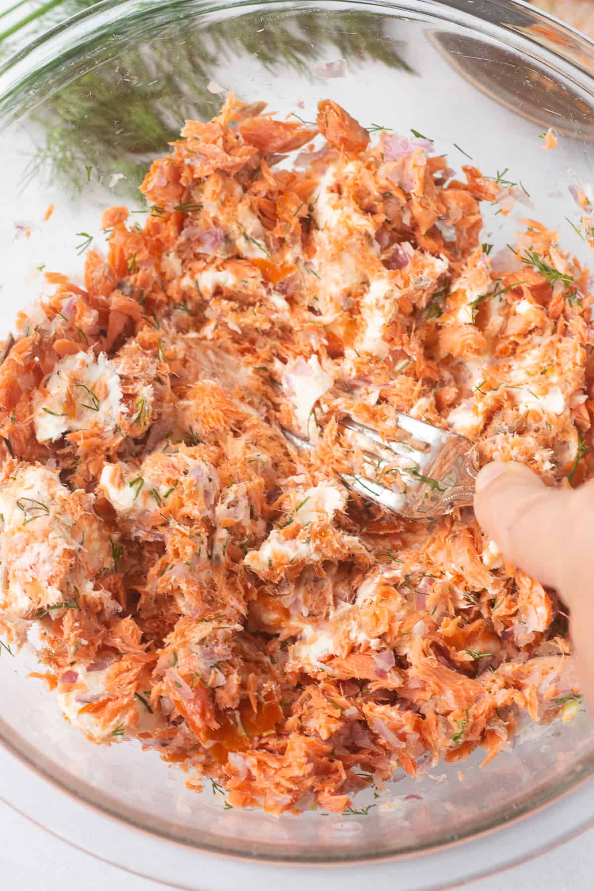 Close up of a fork mashing up ingredients for a salmon ball in a large clear bowl.
