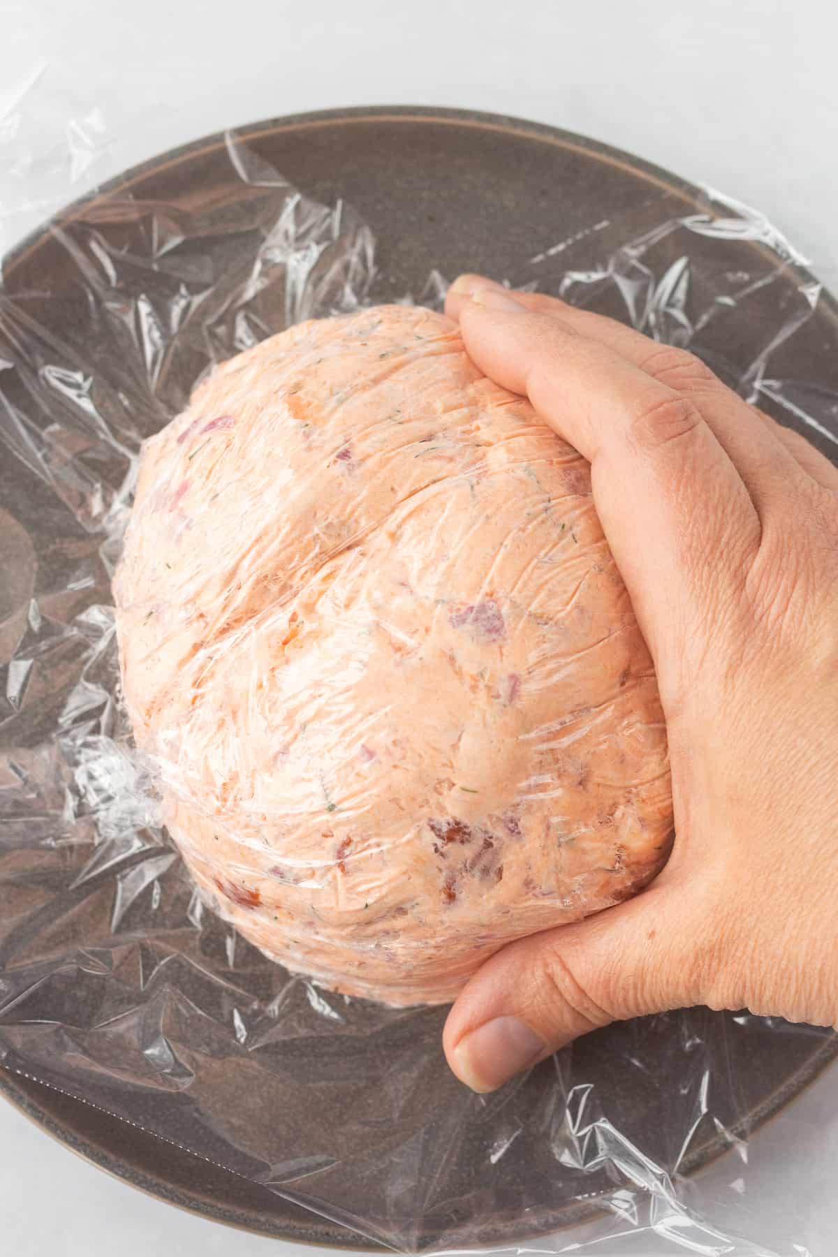 Close up of a hand forming a cream cheese & salmon mixture that's covered in plastic wrap into a ball on a gray plate.