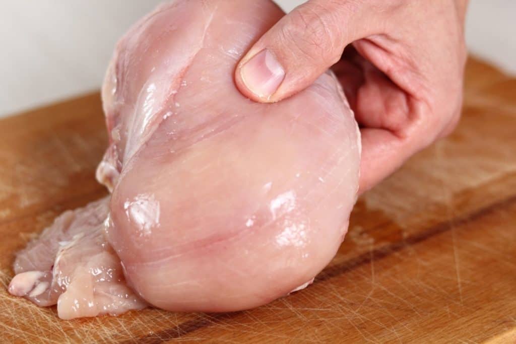 Close up of a hand holding a large raw chicken breast over a wooden cutting board.