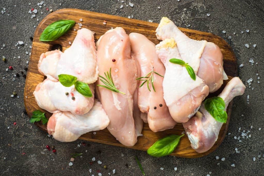 Top down landscape image of raw chicken pieces on a wood cutting board on a dark gray background with basil leaves and salt sprinkled on top.