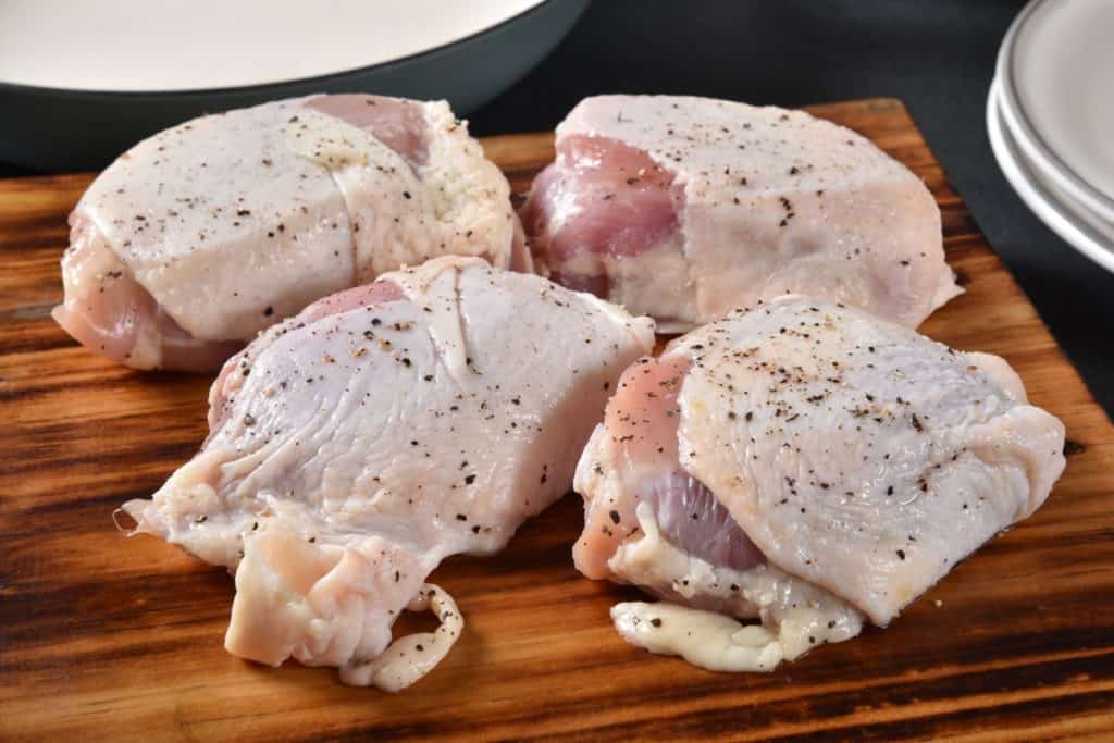 four raw, skin-on, bone-in chicken thighs on a wood cutting board sprinkled with salt and pepper.