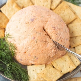 Close up of a salmon cream cheese ball on a gray plate with square crackers around it and a small knife sticking in on the right side.