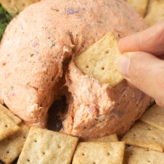 A hand dipping a square cracker into a salmon ball on a gray plate that's surrounded by other square crackers.