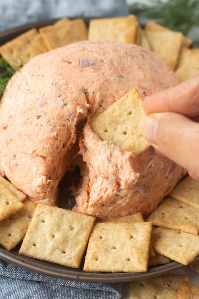 A hand dipping a square cracker into a salmon ball on a gray plate that's surrounded by other square crackers.