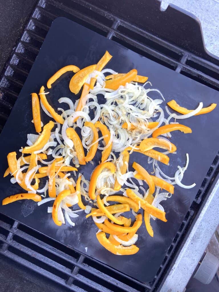 Top down shot of sliced onion and yellow bell peppers on a grill mat, being cooked on a grill.