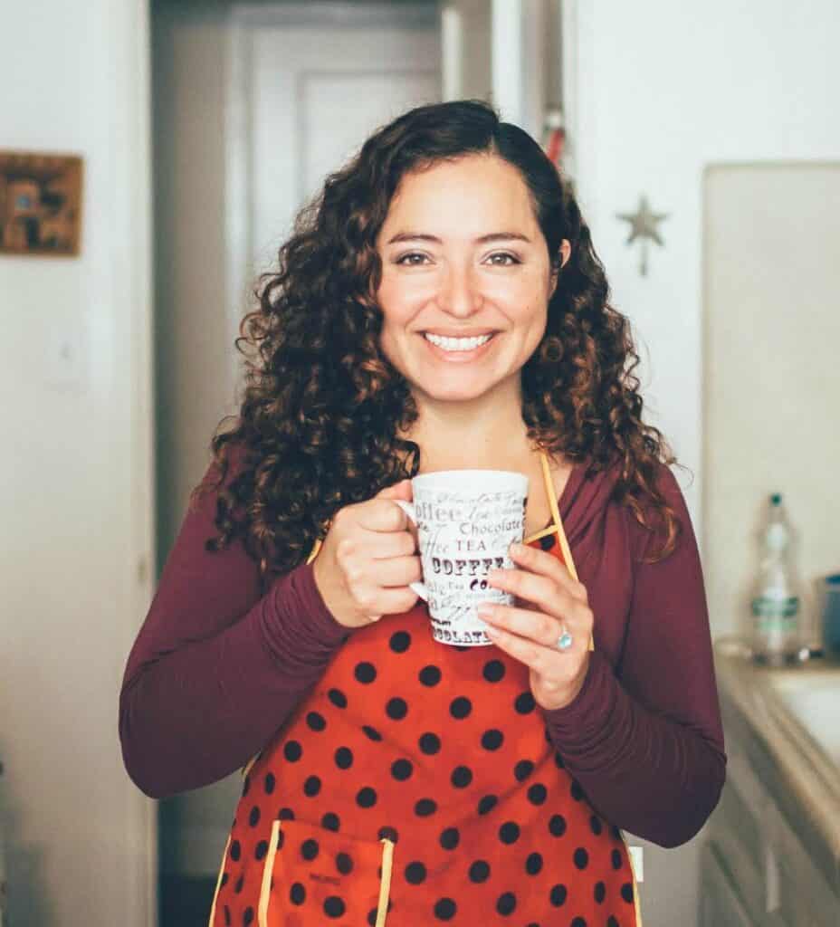 A middle-aged woman with long curly dark hair stands in a kitchen wearing a white and red polka dot apron and holding a white mug. She's smiling at the camer.
