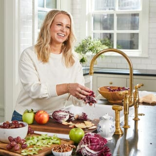 A blonde woman in a white shirt standing rinsing food off in the sink in a white kitchen.