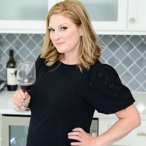 A blonde woman wearing a black shortsleeve shirt stands in a white kitchen holding a glass of red wine.