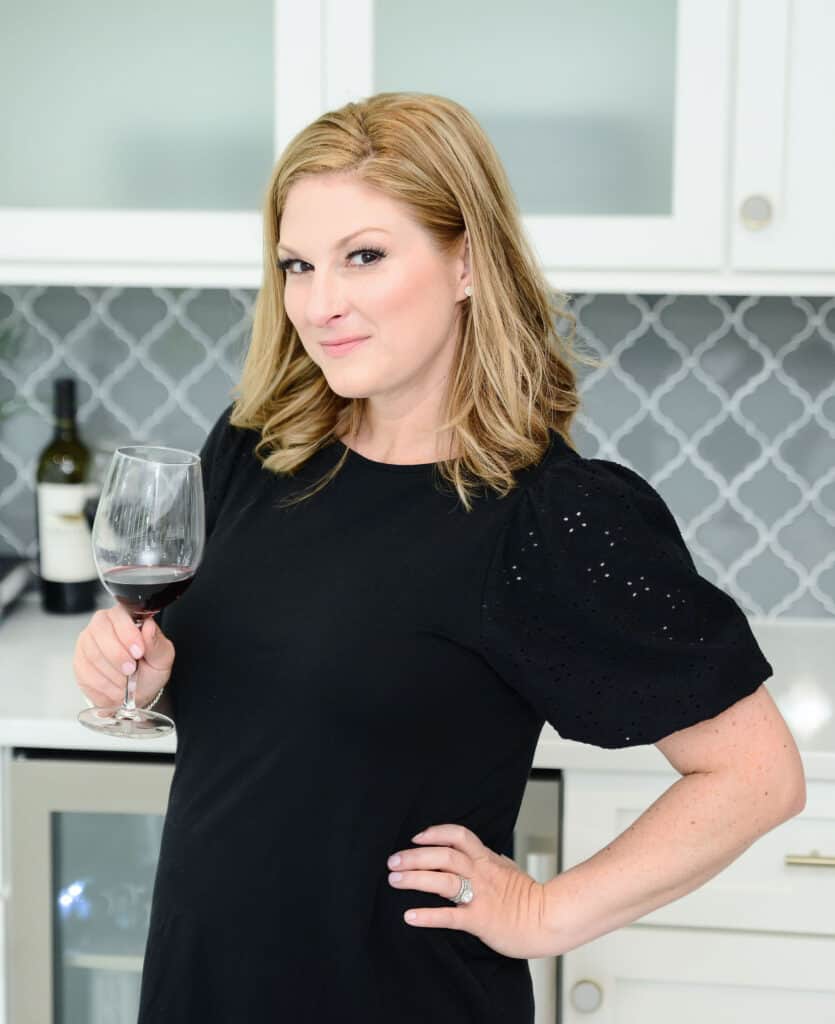 A blonde woman wearing a black shortsleeve shirt stands in a white kitchen holding a glass of red wine.