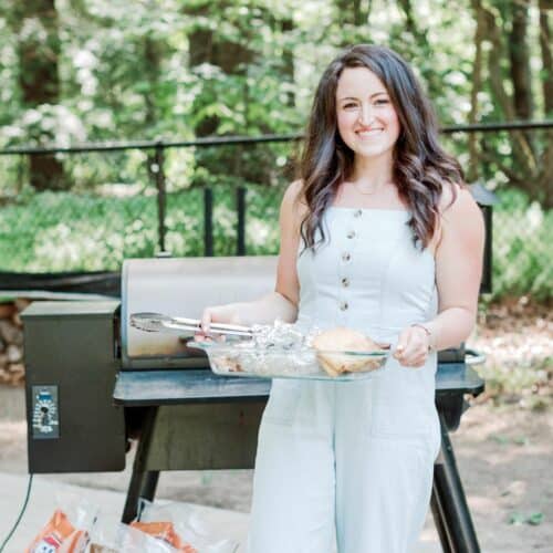 A young woman outside next to a Traeger grill holding food in a platter.