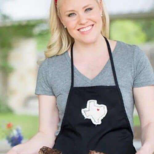 A blonde woman in a gray shirt and black apron holding grilled steak on a cutting board.