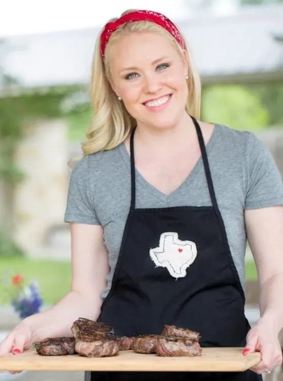 A blonde woman in a gray shirt and black apron holding grilled steak on a cutting board.